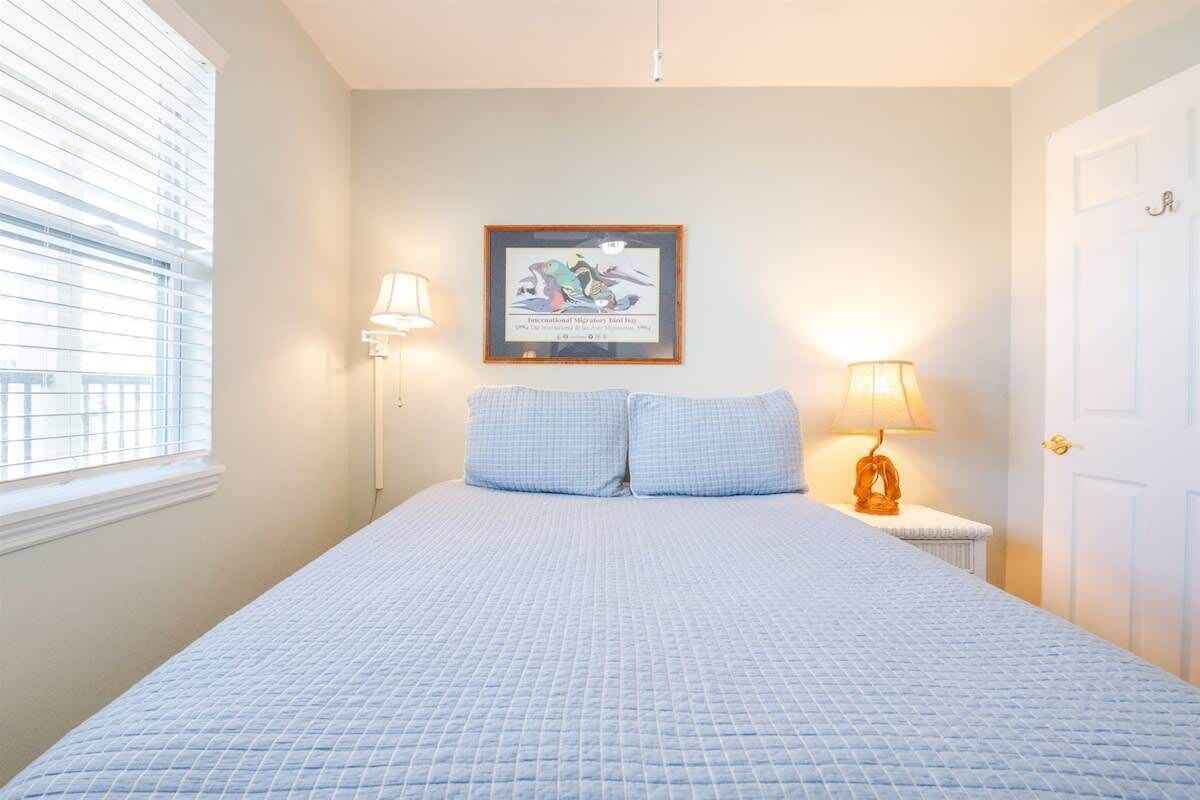 A bedroom with a large bed, two bedside lamps, a framed picture on the wall, and a window with blinds.