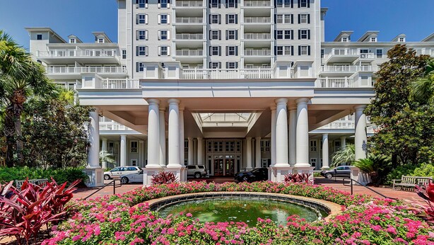 Un gran hotel con entrada central, varios balcones y una fuente rodeada de flores rosadas.