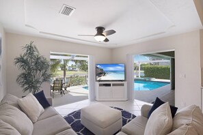 A living room with a television, a ceiling fan, a large plant, and a view of a swimming pool.