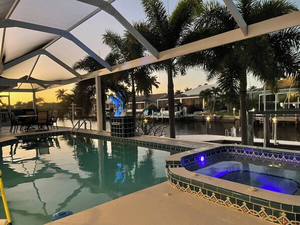 An outdoor swimming pool with a hot tub, surrounded by palm trees and a covered area.