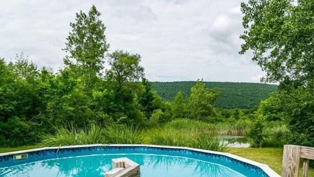 A swimming pool with a wooden deck, surrounded by lush greenery and a hilly landscape in the background.