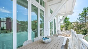 A wooden deck with a table and chairs, large glass windows, and a white railing. - Walk to Seaside! Luxury 6BR w/ Private Pool, Golf Cart & Elevator | Sleeps 20 (Santa Rosa Beach)
