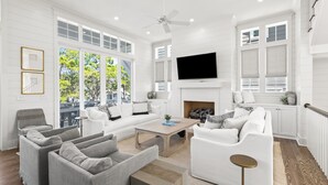 A spacious living room with a white color scheme, large windows, a fireplace, and a flat-screen TV mounted above it. - Walk to Seaside! Luxury 6BR w/ Private Pool, Golf Cart & Elevator | Sleeps 20 (Santa Rosa Beach)