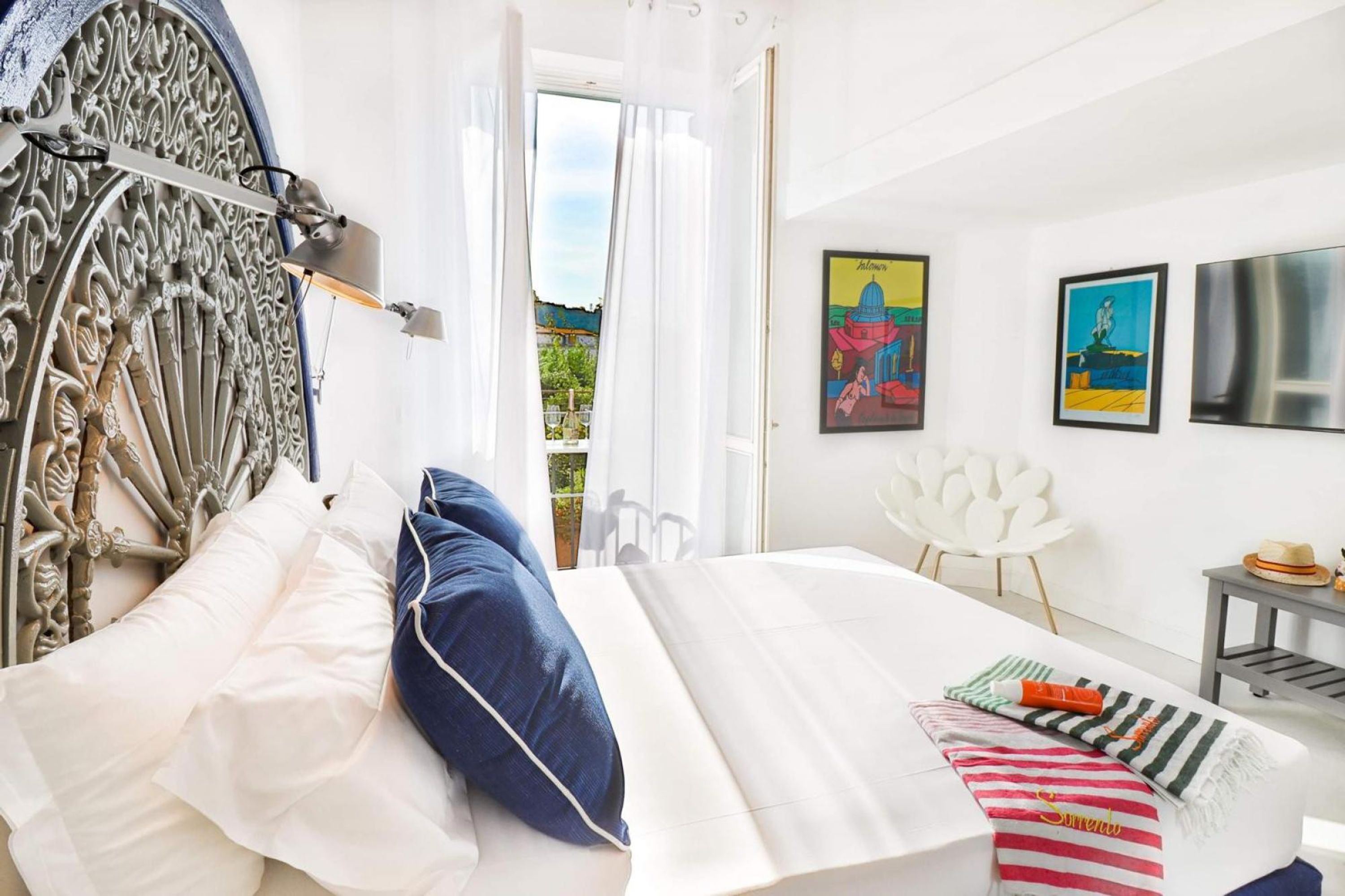 A bedroom with a bed, a white chair, a small table, and artwork on the wall.