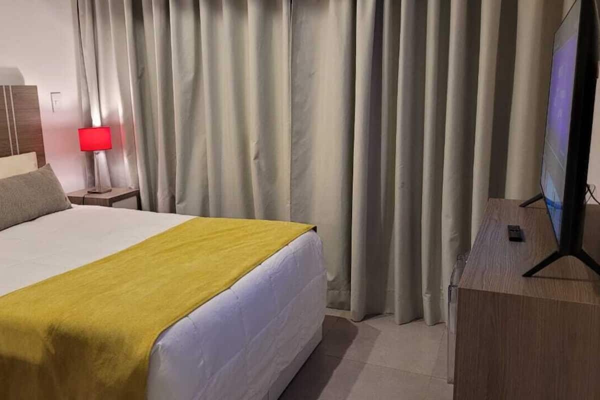 A hotel room with a bed, a TV on a wooden stand, a nightstand with a red lamp, and a curtain.