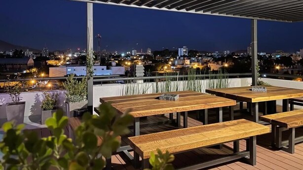 An outdoor patio area with wooden tables and benches, a pergola overhead, and a cityscape view in the background. - Be Haus Temporary Apartments (San Luis)