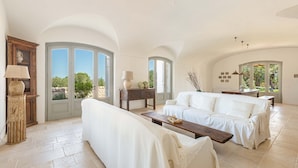 Un salón espacioso con un sofá blanco, una mesa de centro de madera y una lámpara. Amplias ventanas con vistas a zonas verdes.
