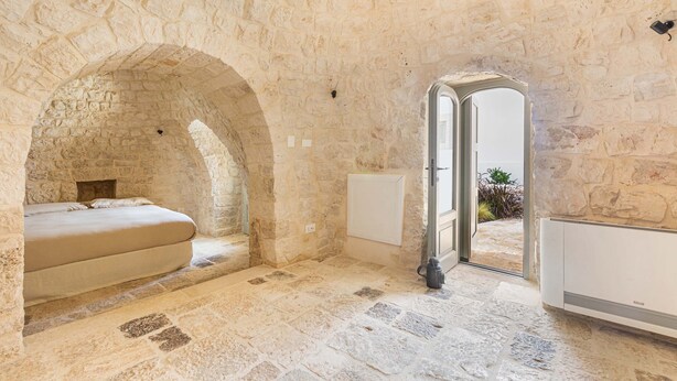 Una habitación con paredes de piedra, una cama, un radiador y un umbral abierto hacia el exterior.