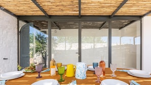 Een eettafel gedekt met borden, bestek en glazen, onder een houten pergola met glazen deuren.