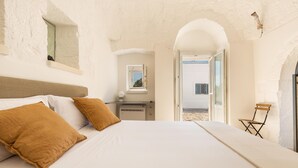 Een slaapkamer met een groot bed, een klein bureau en een stoel. Er is een raam en een boogvormige doorgang naar buiten.