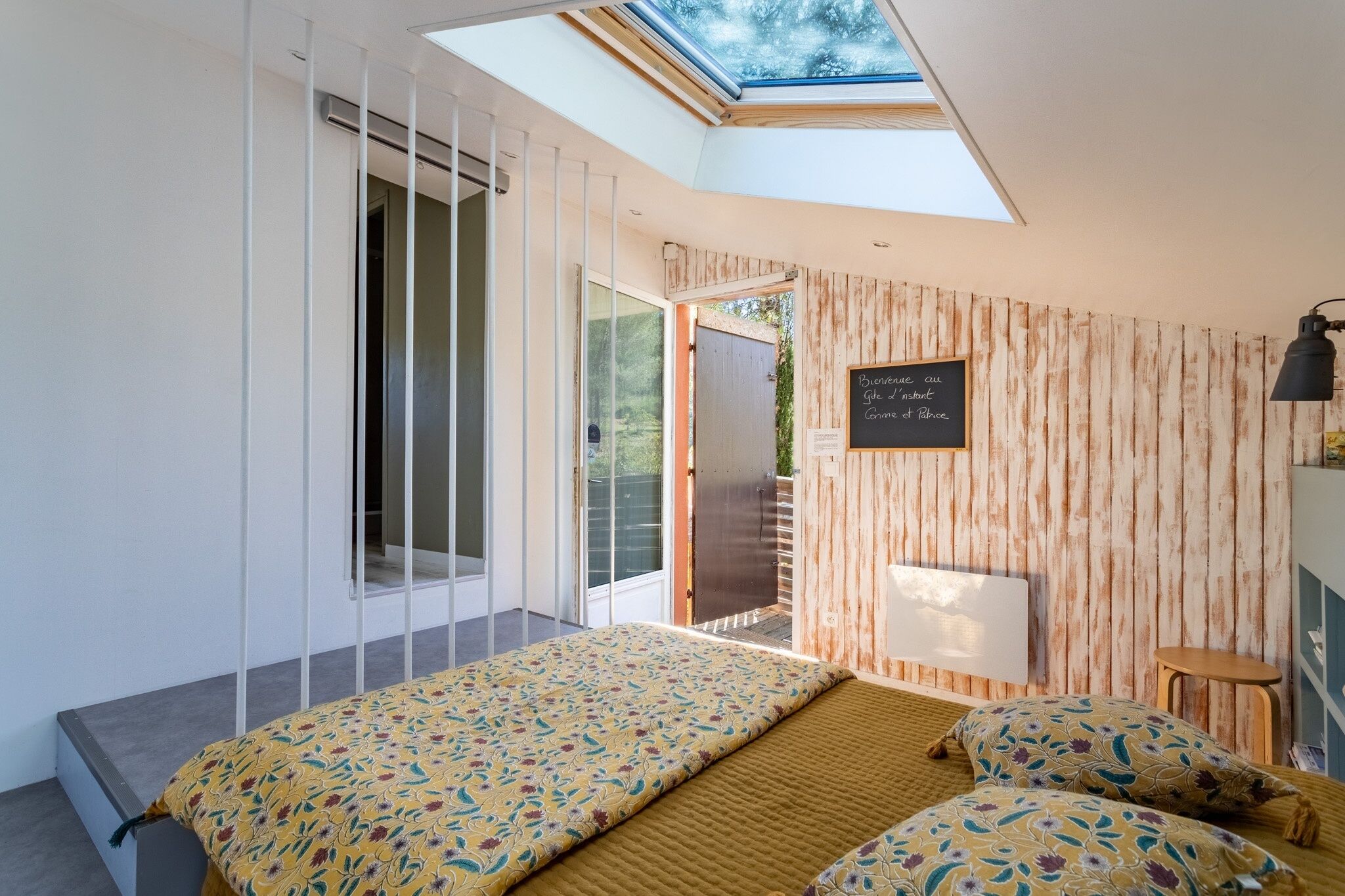 A bedroom with a skylight, a bed with patterned bedding, a wooden wall, and a chalkboard with text.