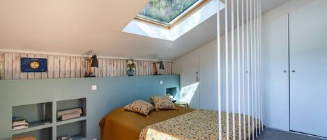 A bedroom with a skylight, a bed with patterned bedding, a bedside table, a shelf unit, and a wardrobe.