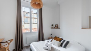 A hotel room with a bed, a chair, a nightstand, a window with curtains, and a view of buildings outside.