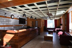 A reception area with a wooden reception desk, a clock, and a television. There is a seating area with a sofa and a table. - Bradhi - Riverside Resort Jibhi (Banjar)