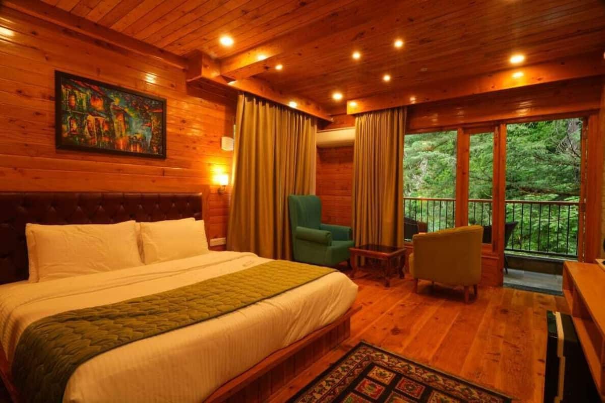 A wooden cabin-style room with a bed, two chairs, a small table, and a balcony with a view of the forest.