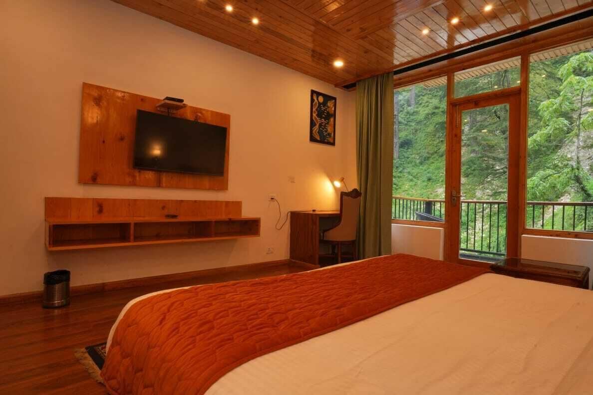 A bedroom with a large bed, a TV mounted on the wall, a desk, and a window with a view of greenery.