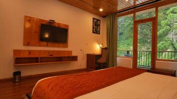 A bedroom with a large bed, a TV mounted on the wall, a desk, and a window with a view of greenery.