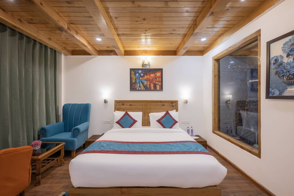 A hotel room with a wooden ceiling, a large bed with a blue and red bedspread, a blue armchair, a small table with a vase, and a painting on the wall.