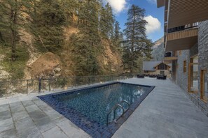 A pool area with stone flooring, a glass railing, and a view of a mountainous forest. - Bradhi - Riverside Resort Jibhi (Banjar)