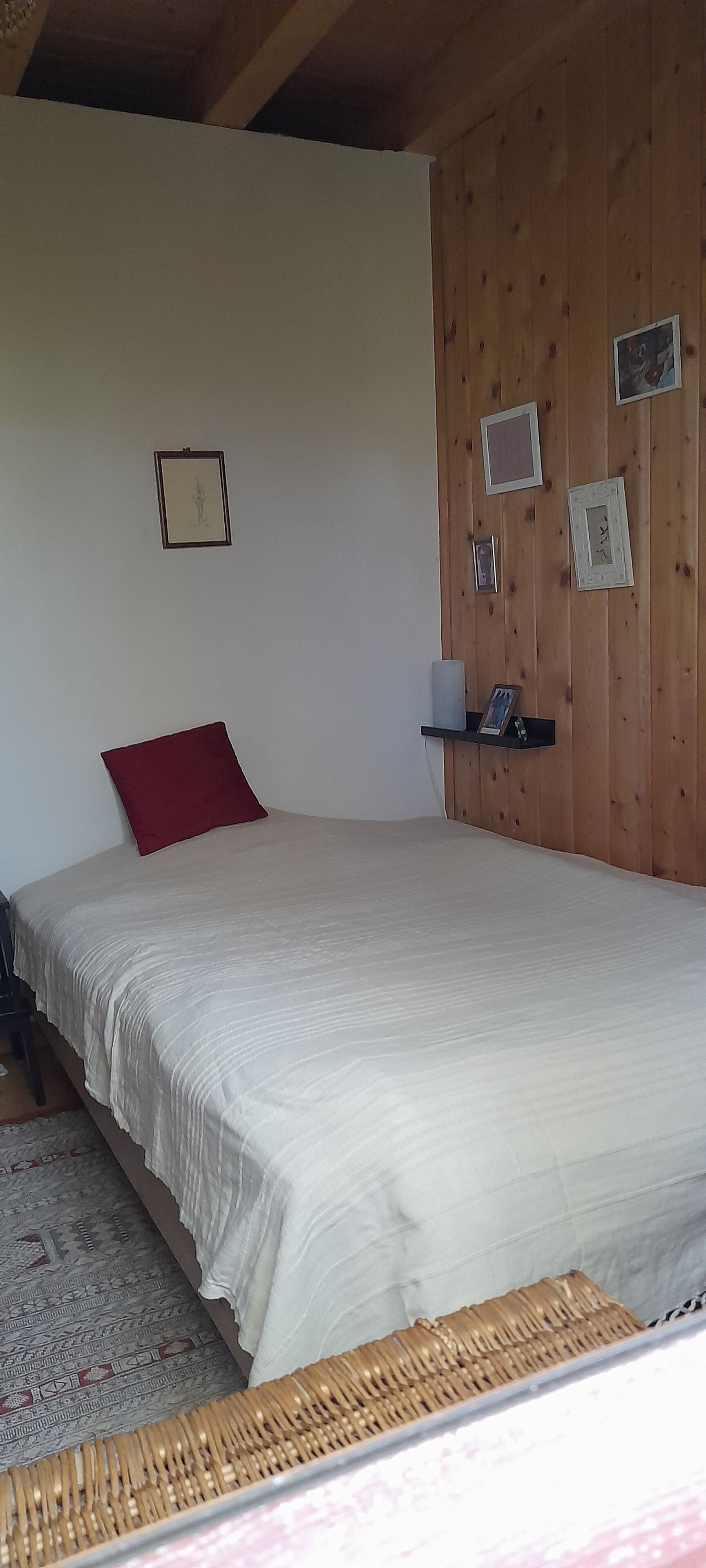 A bedroom with a large bed, a red pillow, a wooden wall, and a shelf with framed pictures.