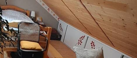 Une chambre mansardée chaleureuse avec un plafond en pente en bois, un lit avec une literie à motifs floraux, une chaise et une petite table.