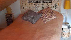 Una cama con colcha color melocotón y dos almohadas, cabecera de madera, un gráfico botánico en la pared y una lámpara sobre una mesita de noche.