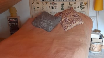 Uma cama com colcha cor de pêssego e duas almofadas, cabeceira de madeira, um diagrama botânico na parede e uma luminária em uma mesinha de cabeceira.