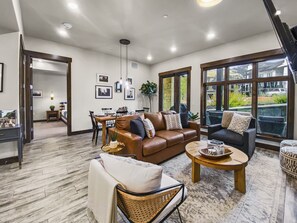 Amplia sala de estar con un sofá de cuero marrón, un sofá gris oscuro y un sillón de mimbre. Hay una zona de comedor con mesa y sillas, y ventanas grandes con vistas al exterior.