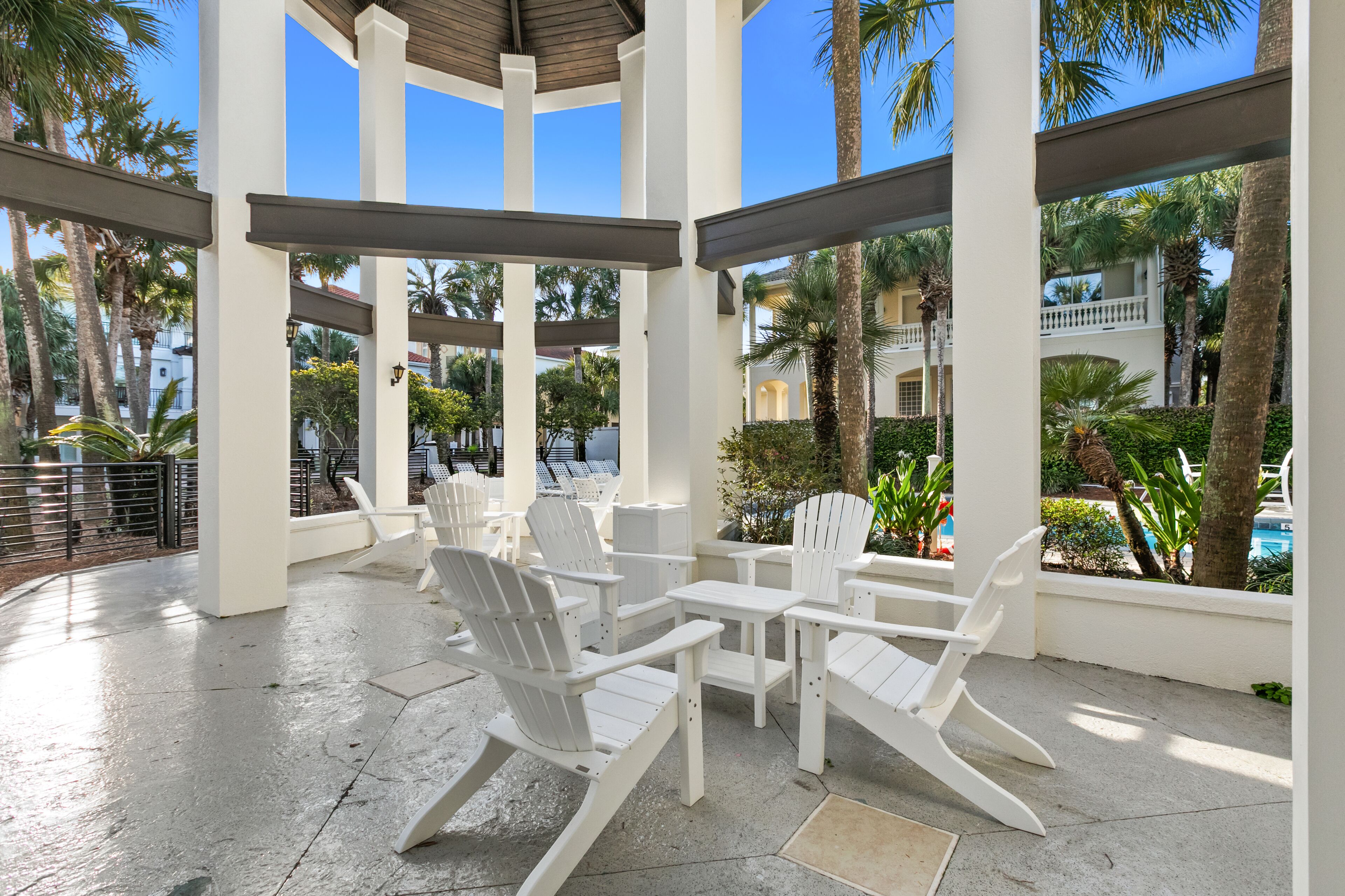 A covered outdoor seating area with white chairs and tables, surrounded by palm trees and a pool.