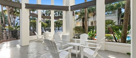 A covered outdoor seating area with white chairs and tables, surrounded by palm trees and a pool.