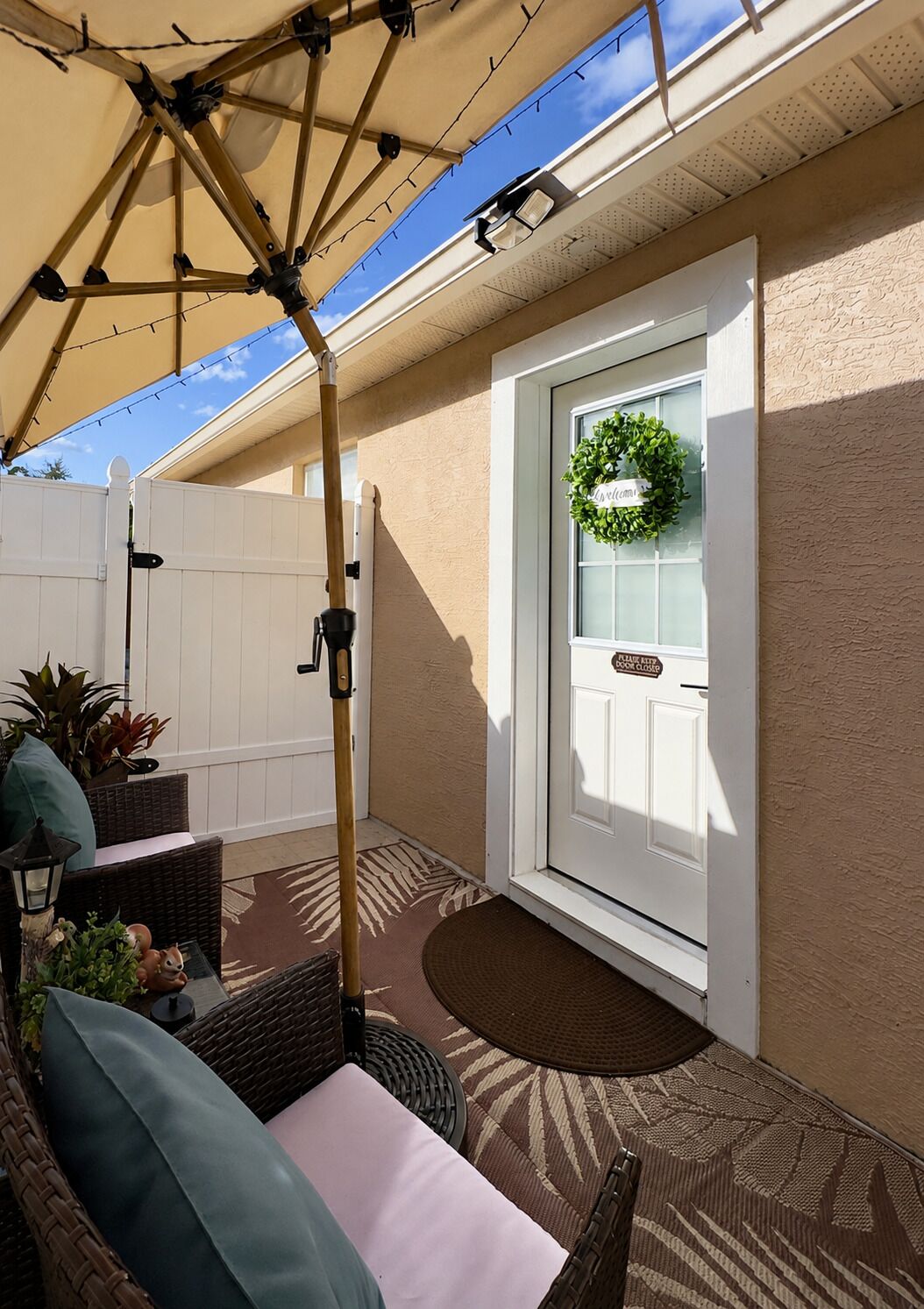 A covered patio area with a wicker sofa, a small table with a lamp, and a welcome wreath on the door.