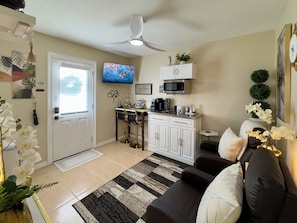 A living room with a sofa, a coffee table, a TV, a ceiling fan, and a kitchen area with a microwave and cabinets.