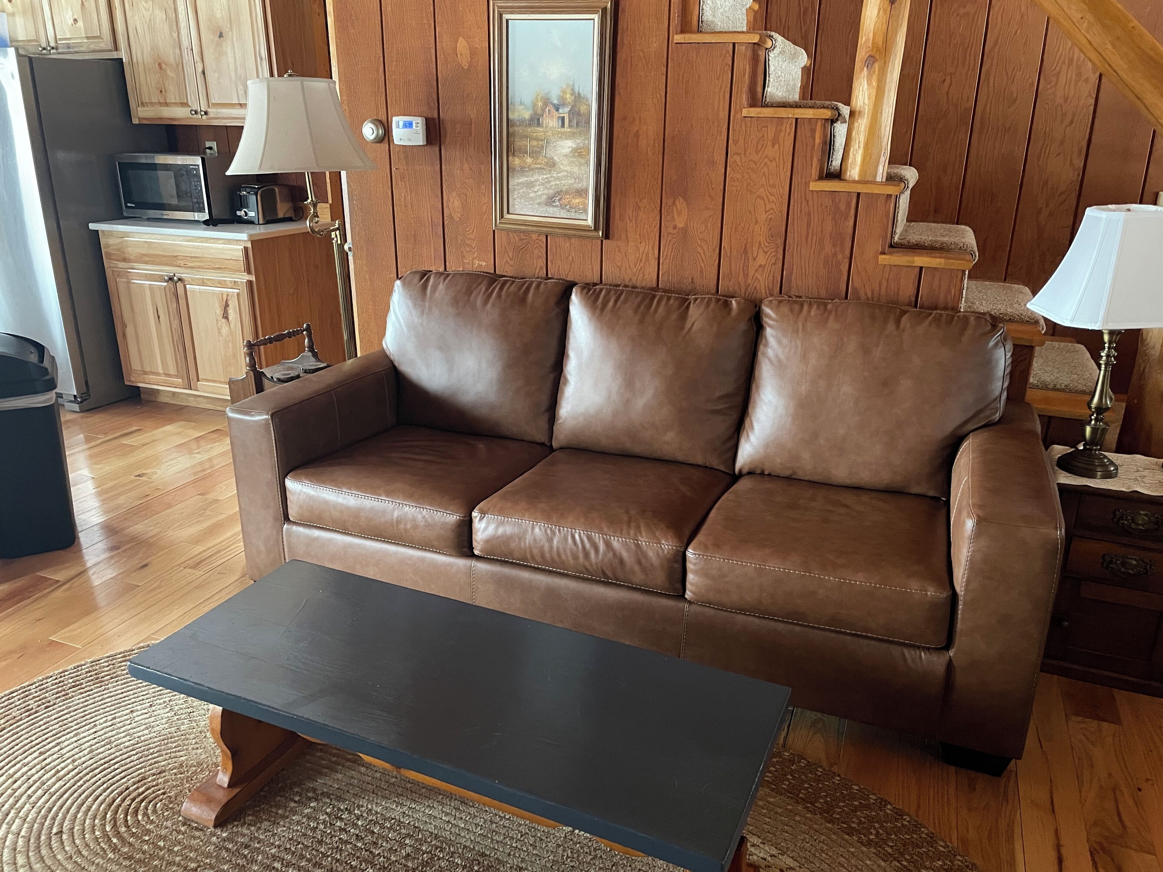 Un salon chaleureux avec un canapé en cuir marron, une table basse noire, un plancher en bois, un coin cuisine doté de meubles en bois, un four à micro-ondes et un tableau accroché au mur.