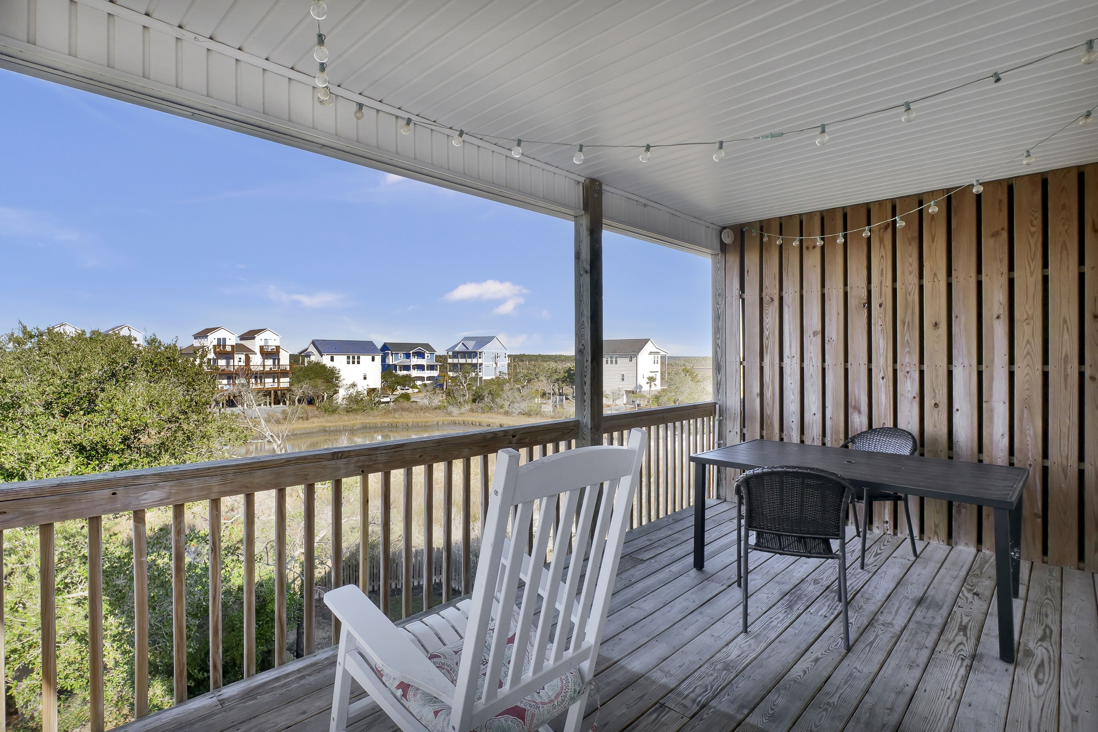 A covered outdoor area with a wooden deck, a white chair, a table, and a view of houses across a body of water.