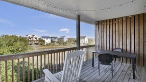 A covered outdoor area with a wooden deck, a white chair, a table, and a view of houses across a body of water. - Sunset Horizon | Private Fenced-in Backyard | Surf City! (surf ctiy)