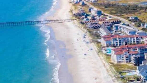 An aerial view of a beach with a pier extending into the water, a row of houses along the shore, and a clear sky. - Sunset Horizon | Private Fenced-in Backyard | Surf City! (surf ctiy)