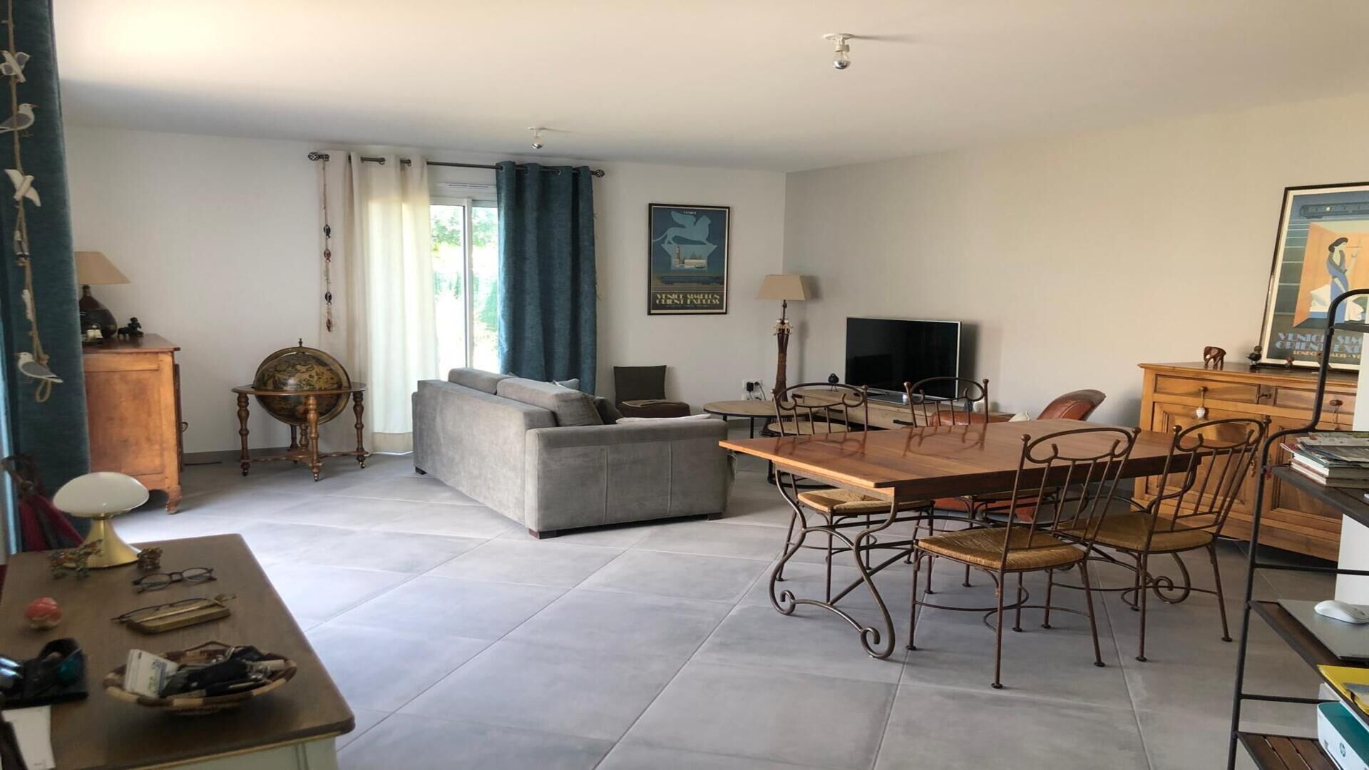 A spacious living room with a grey sofa, a wooden dining table with chairs, a television, and a globe on a side table.