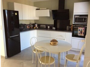 A compact kitchen with white cabinets, a black refrigerator, a microwave, and a toaster. There is a round table with four chairs and a basket of bread on it. - Commentaire Sous peu (L'Houmeau)