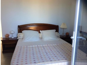 A bedroom with a wooden headboard, two white pillows, a patterned bedspread, a nightstand with a lamp, and a window with a view. - Commentaire Sous peu (L'Houmeau)