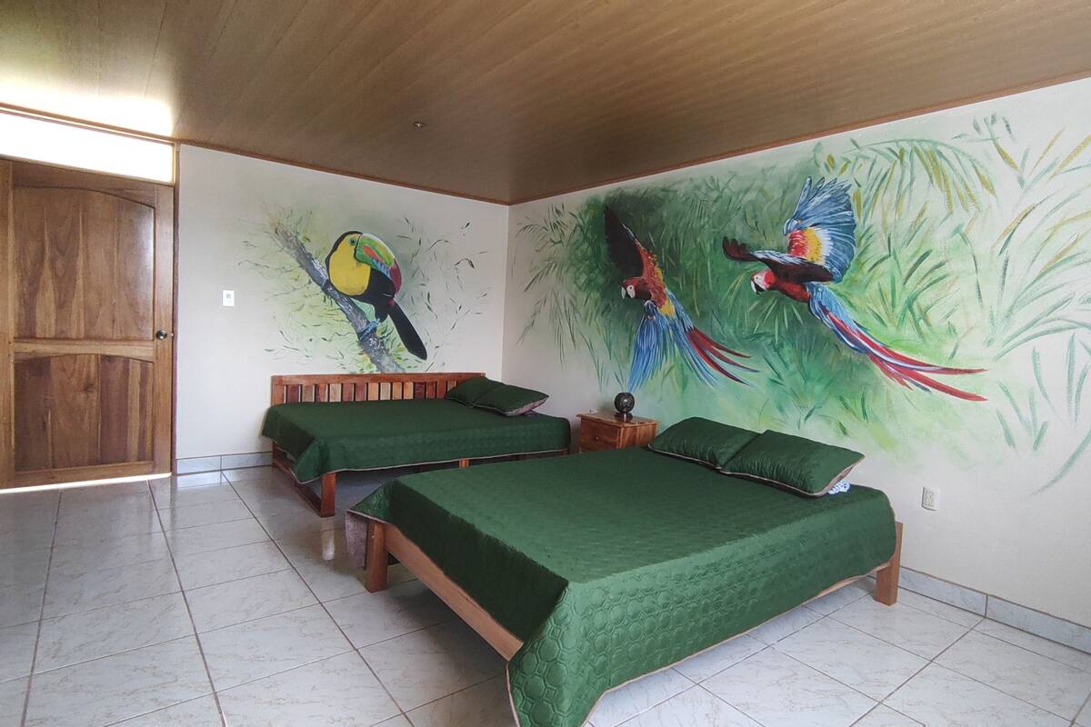 Une pièce avec deux lits en bois, une couverture verte, une porte en bois et une fresque représentant des oiseaux aux couleurs vives sur le mur.