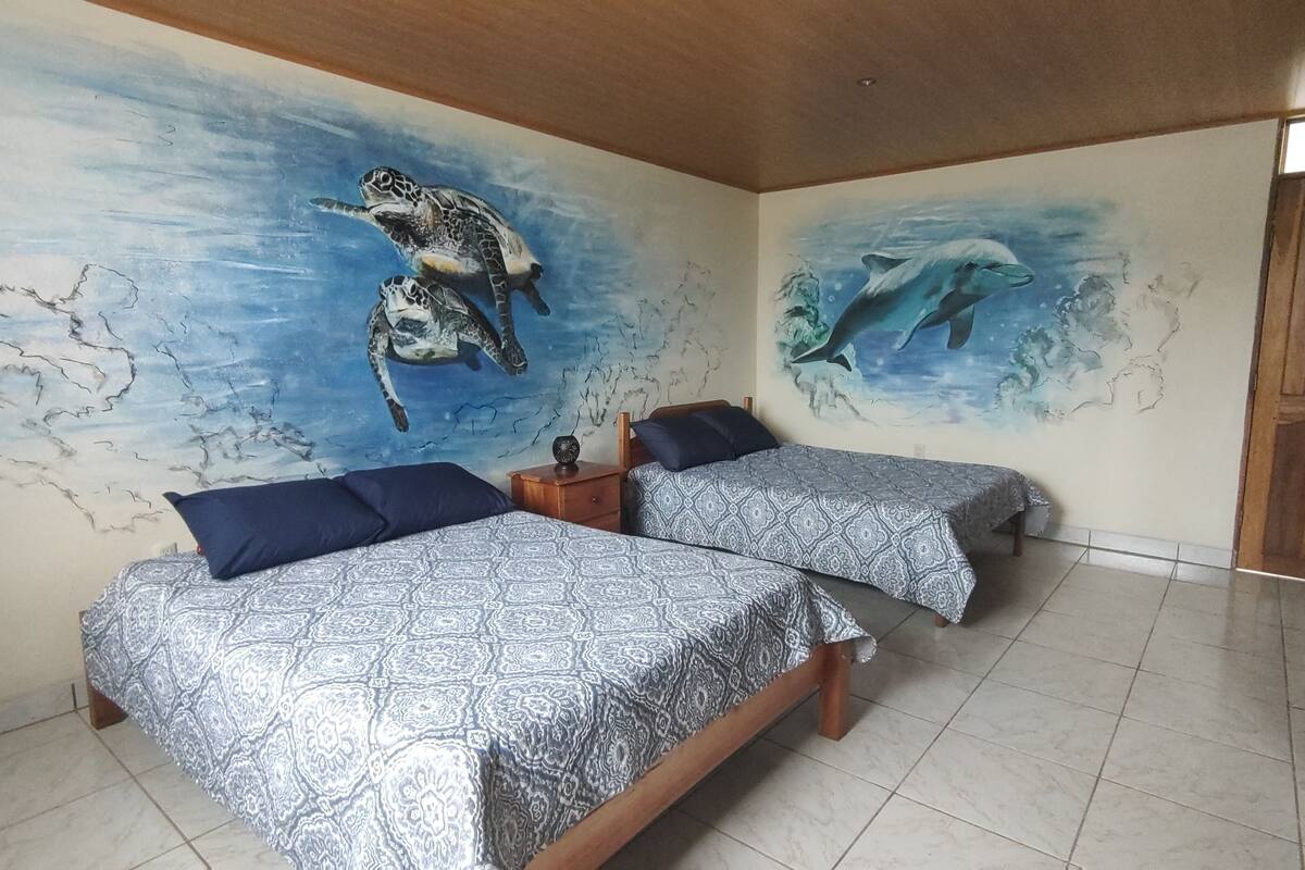 Une chambre avec deux lits, chacun agrémenté de draps à motifs et d’oreillers bleus, et un mur décoré d’une fresque représentant des tortues de mer et des dauphins.