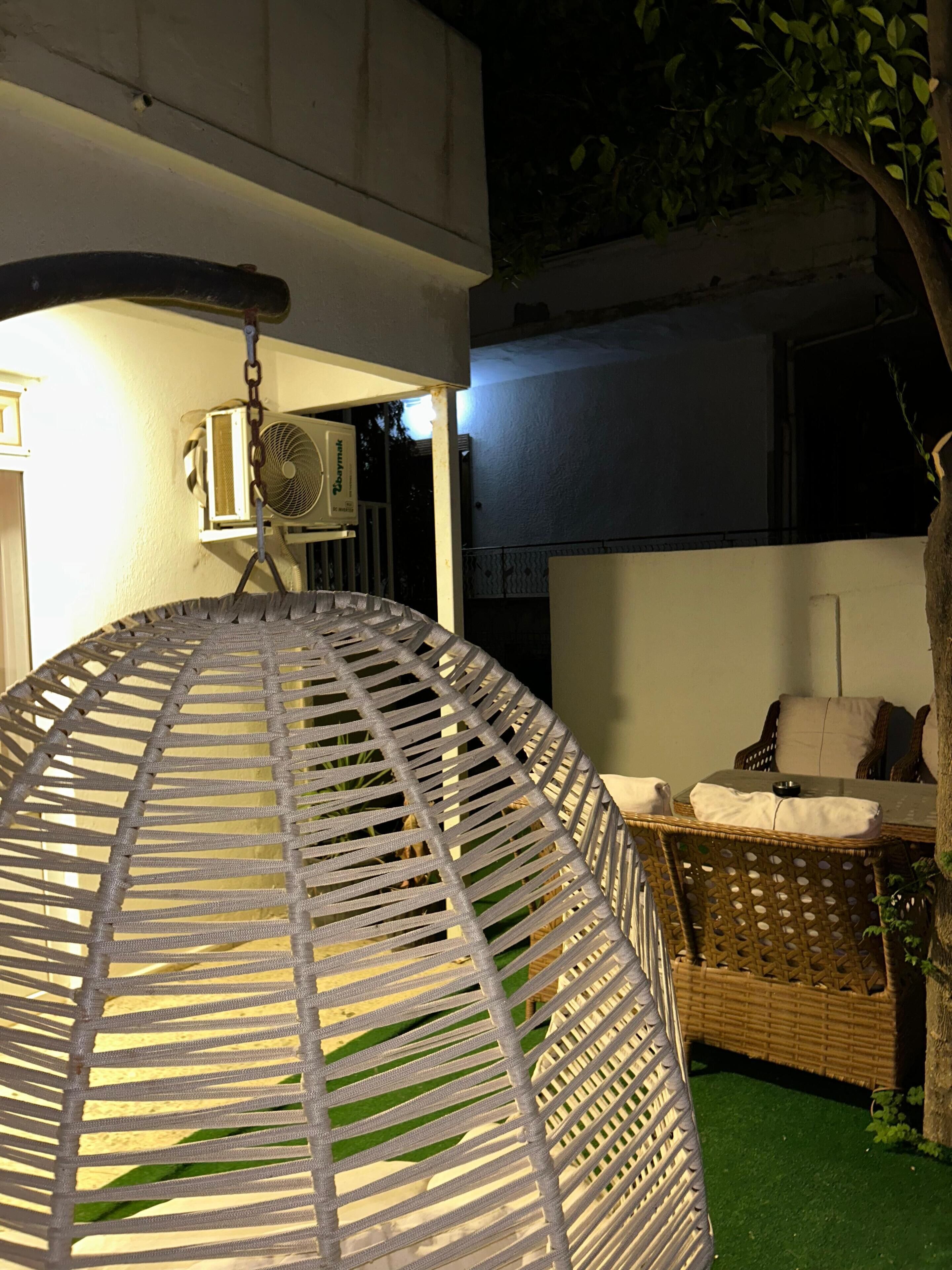 A circular woven structure, possibly a hanging chair, in an outdoor setting with a wicker sofa and cushions, an air conditioning unit, and a tree.