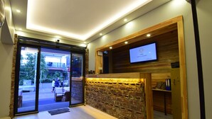 Zona de recepción de un hotel, con un mostrador de madera, una pantalla de televisión y un puerta de cristal que da al exterior.
