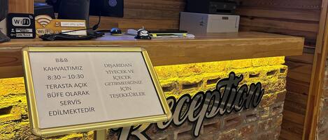 Egy recepciós pult, melynek háttérfalát fából készítették, és amelyen feltüntették a recepción nyitva tartási időt.