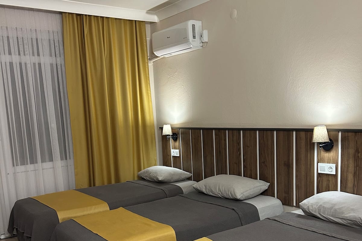 A hotel room with two beds, a wooden floor, a window with curtains, an air conditioning unit, and wall-mounted lamps.