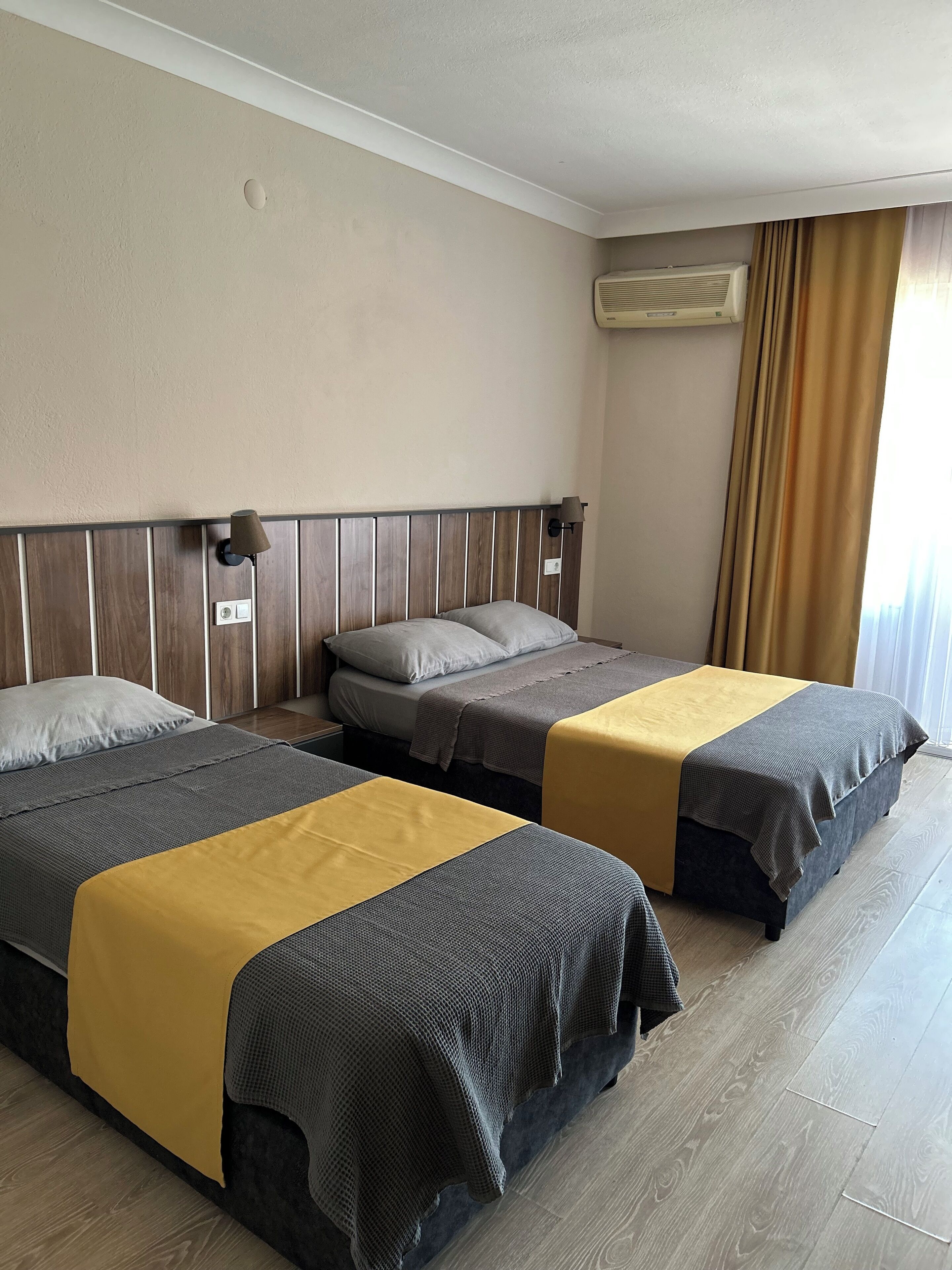 A hotel room with two beds, a wooden headboard, a wall-mounted air conditioner, and a window with curtains.