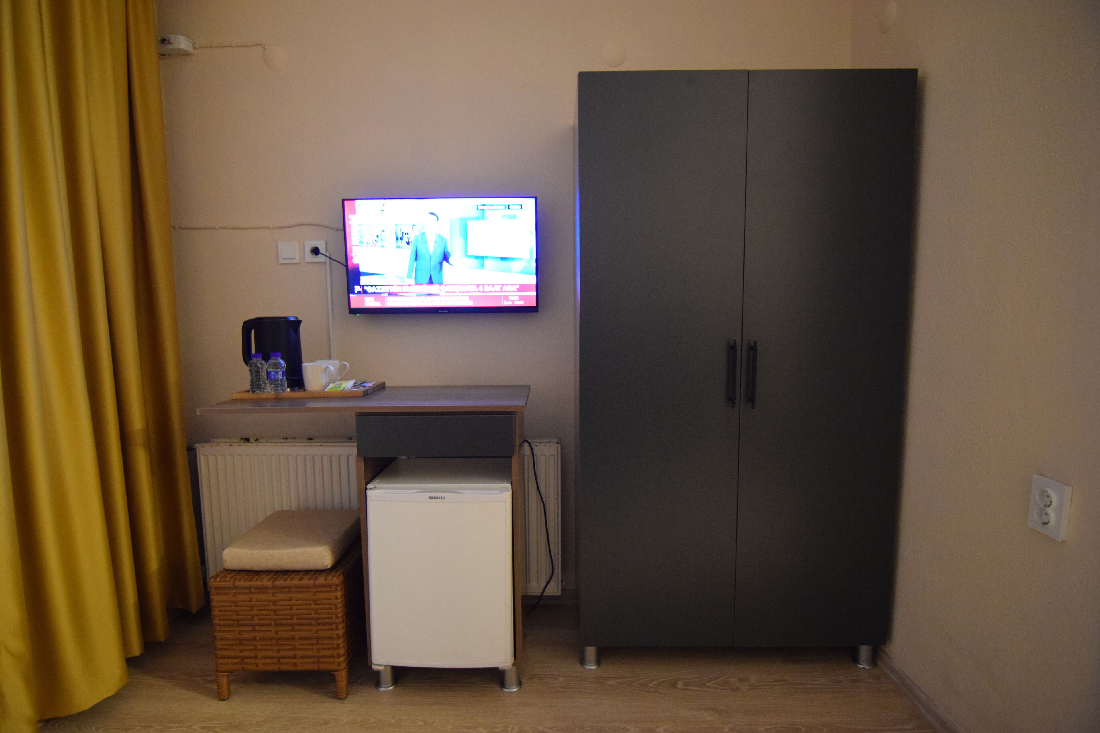 A room with a television mounted on the wall, a small white refrigerator, a dark wardrobe, a wicker bench, and a radiator.