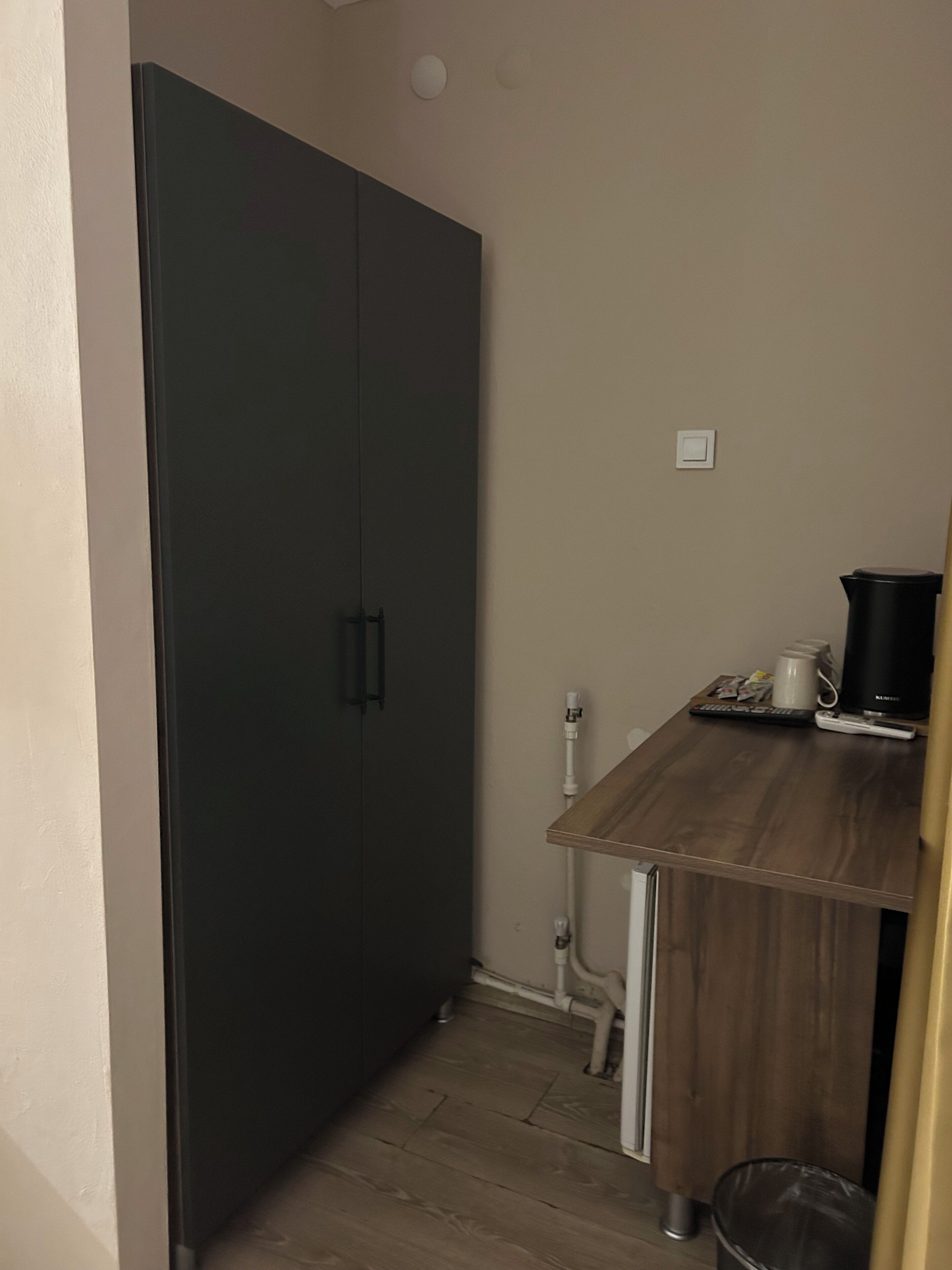 A small room with a dark wardrobe, a wooden desk with a coffee maker, and a trash bin.