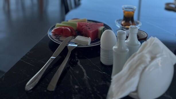 Una mesa de comedor con una porción de sandía y otras frutas, un vaso de jugo de naranja, saleros y pimenteros, y una servilleta. La mesa está colocada al aire libre, con vista al mar y edificios al fondo.