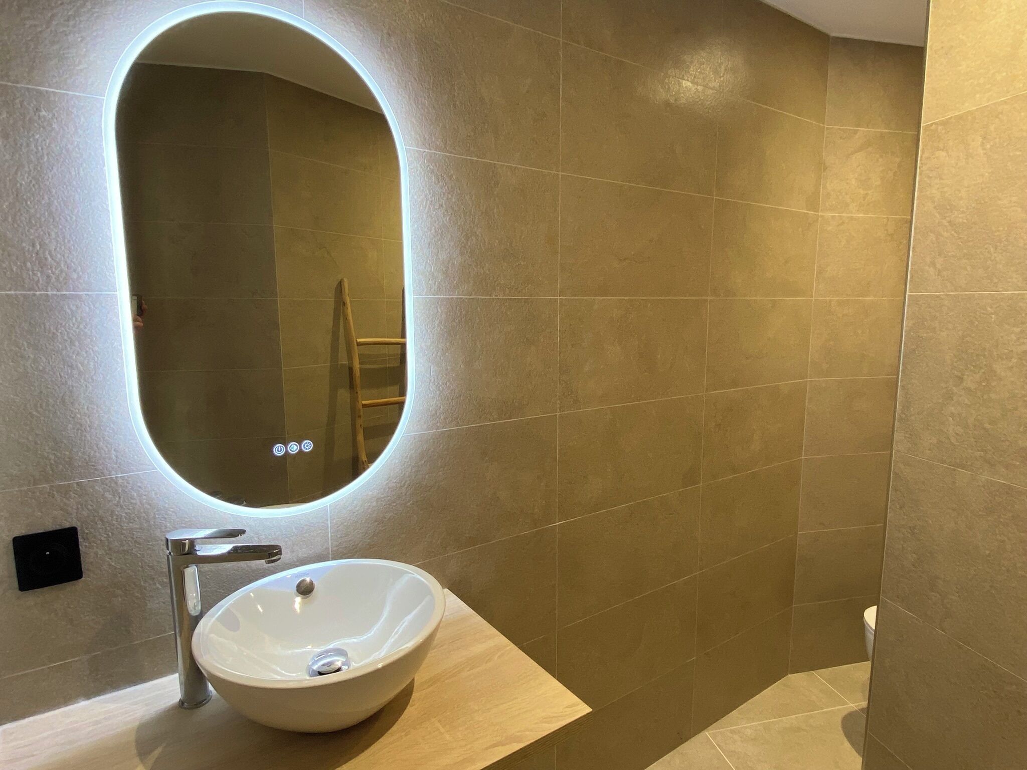 Un baño moderno con un espejo redondo, un lavabo blanco y azulejos color beige.